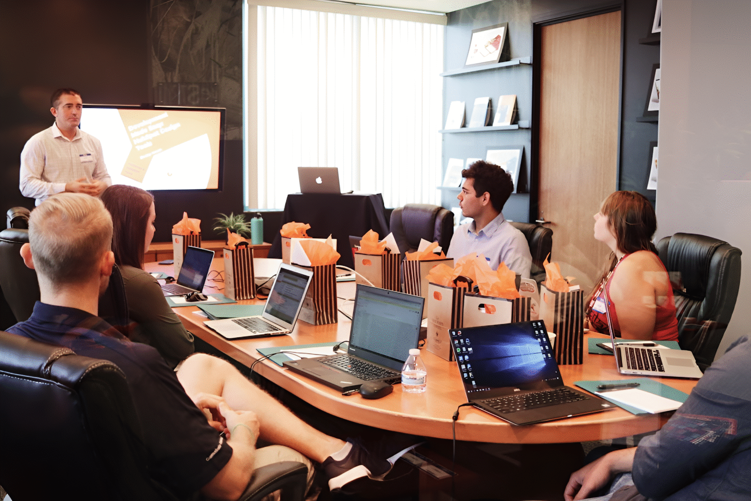 Several individuals are seated at a table with laptops, engaged in a discussion about proposal management techniques.